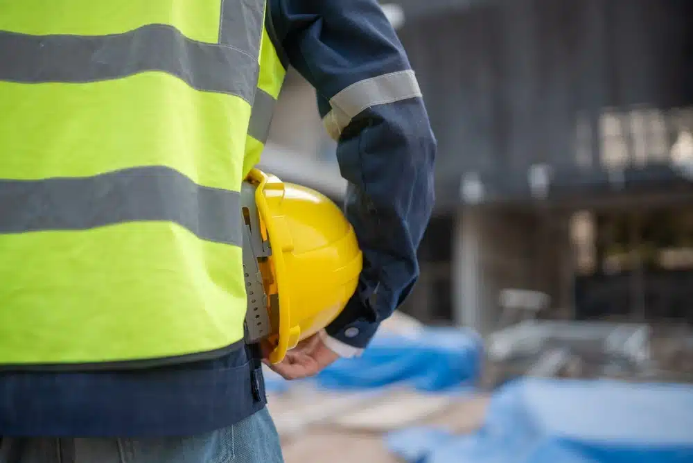 Une personne portant un gilet haute visibilité et une veste bleu foncé tient un casque de chantier jaune sur un site de construction. L'atmosphère reflète la concentration et la préparation.