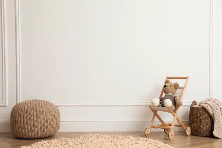 Chambre de bébé chaleureuse avec un pouf en tricot, un landau en bois pour jouets contenant un ours en peluche et un panier tressé avec une couverture, le tout posé sur un parquet en bois. Tons doux et neutres.