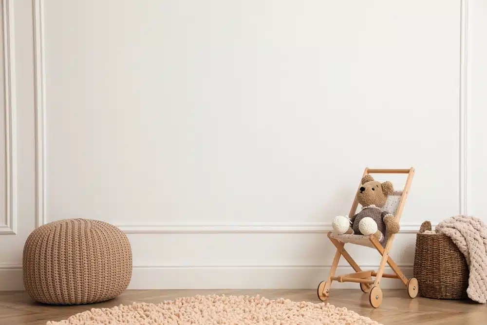 Chambre de bébé chaleureuse avec un pouf en tricot, un landau en bois pour jouets contenant un ours en peluche et un panier tressé avec une couverture, le tout posé sur un parquet en bois. Tons doux et neutres.