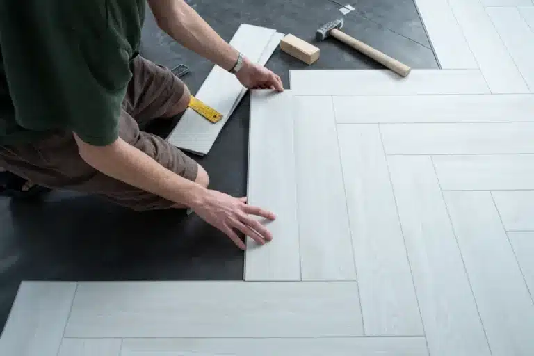 Une personne agenouillée sur un sol sombre pose avec soin des lames de bois clair en motif à chevrons. Des outils tels qu'un marteau et un mètre ruban se trouvent à proximité.