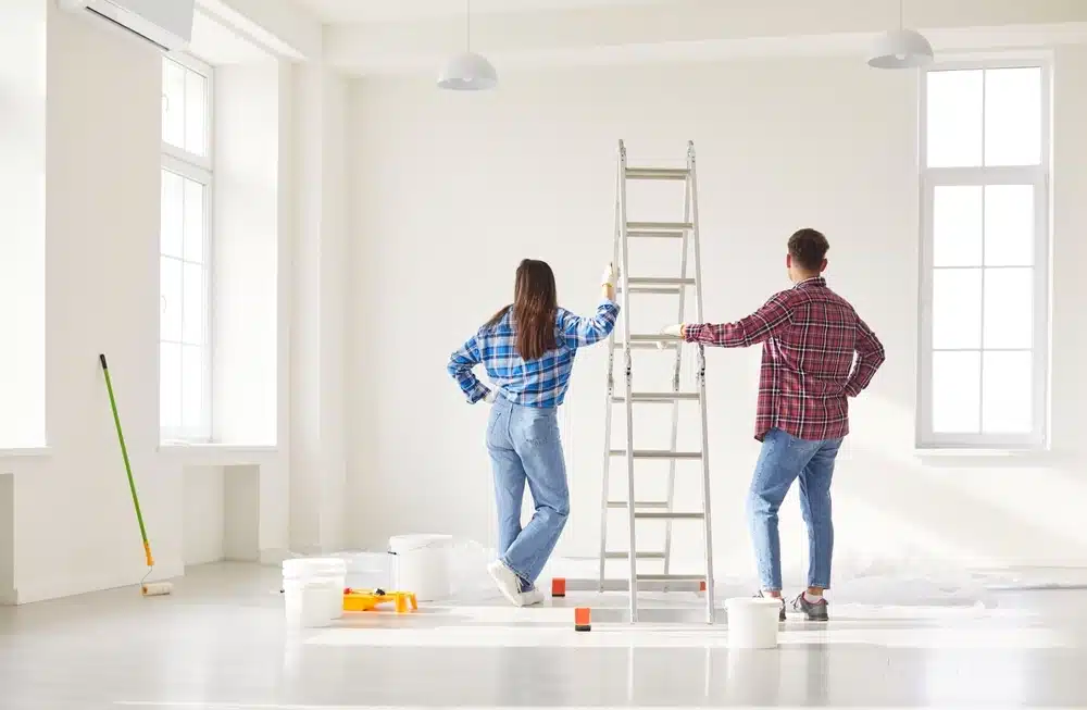 Deux personnes vêtues de chemises à carreaux décontractées se tiennent près d'une échelle dans une pièce lumineuse. Du matériel de peinture est éparpillé aux alentours, suggérant un projet de rénovation.