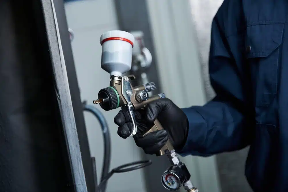 Une personne vêtue d'une tenue de travail bleue et portant des gants noirs tient un pistolet à peinture, en se concentrant sur la précision. Le cadre évoque un environnement industriel ou un atelier.