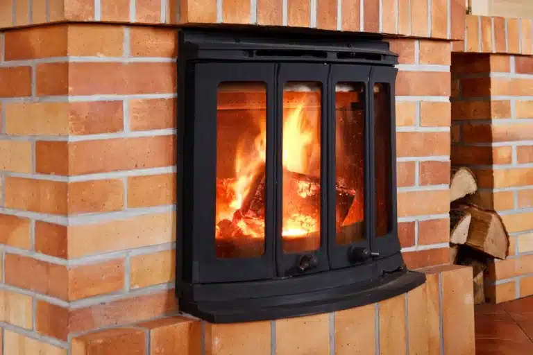 Une cheminée en brique chaleureuse, dotée d'une porte en métal noir, abrite un feu vif et crépitant. Des bûches empilées sont visibles à proximité, créant une atmosphère chaleureuse.