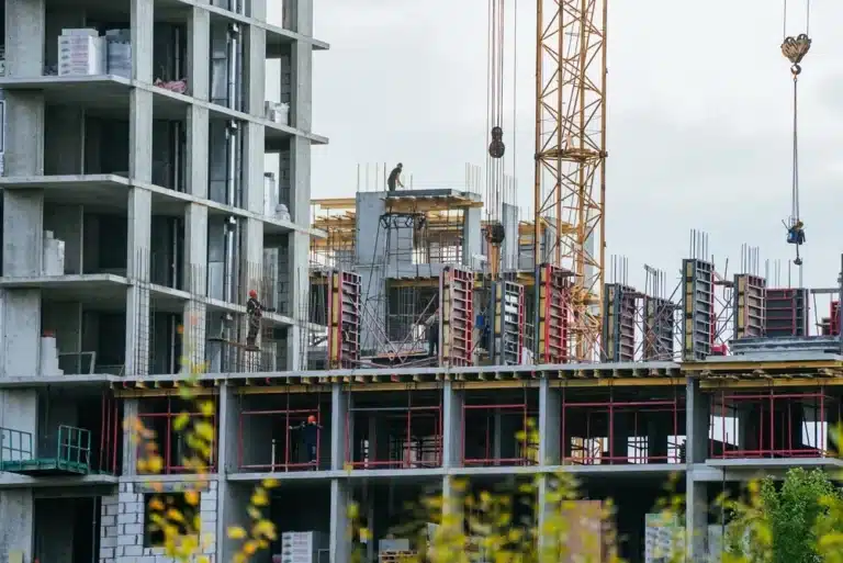 Chantier de construction avec un bâtiment en béton partiellement construit et une grue. Des ouvriers se trouvent sur des échafaudages et du matériel est présent sur le site. Ciel couvert en arrière-plan.
