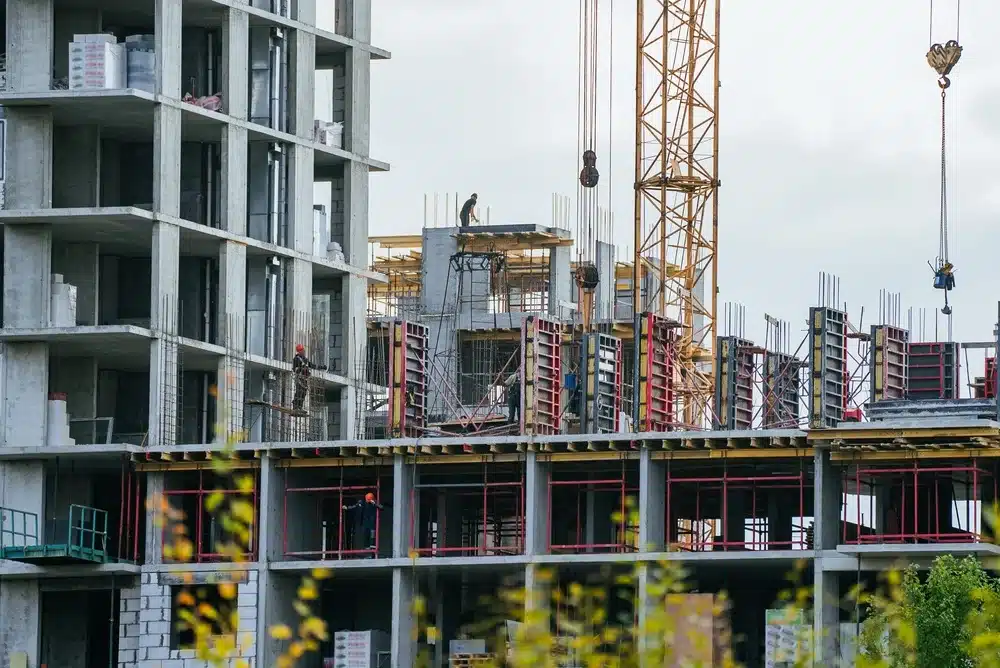 Chantier de construction avec un bâtiment en béton partiellement construit et une grue. Des ouvriers se trouvent sur des échafaudages et du matériel est présent sur le site. Ciel couvert en arrière-plan.