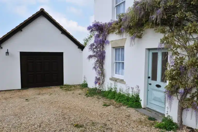 Maison blanche avec une porte bleue et de la glycine violette grimpant sur les murs. À côté se trouve un garage blanc doté d'une porte en bois foncé. Une allée en gravier s'étend devant.