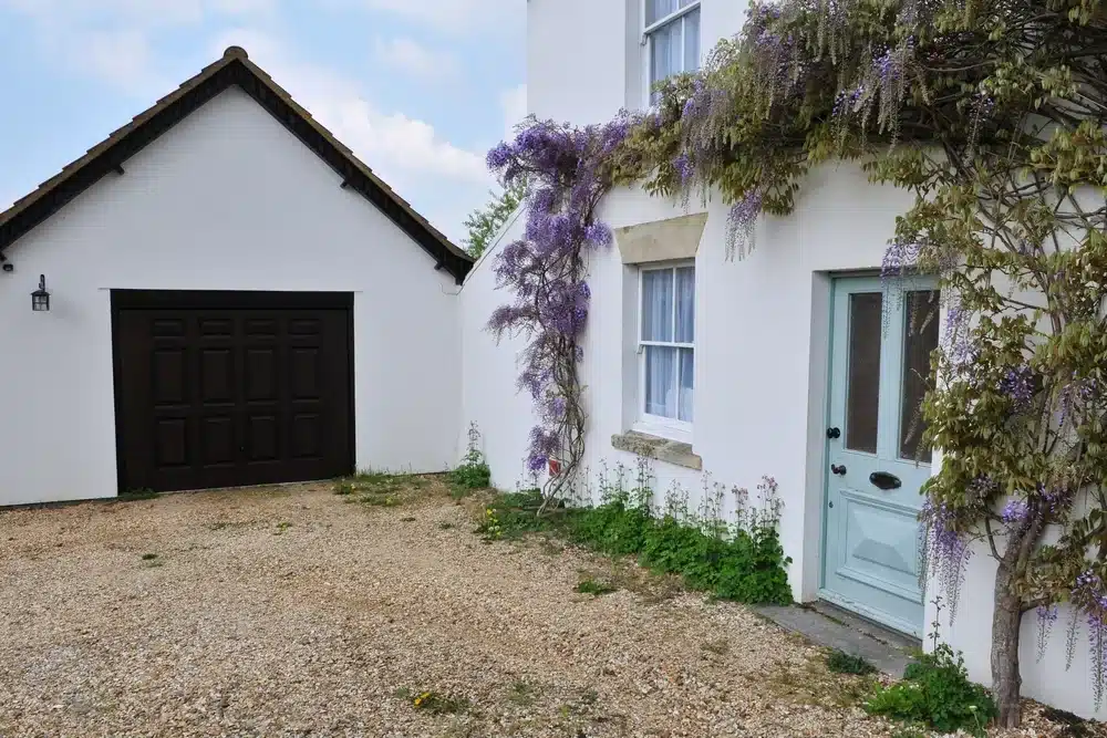 Maison blanche avec une porte bleue et de la glycine violette grimpant sur les murs. À côté se trouve un garage blanc doté d'une porte en bois foncé. Une allée en gravier s'étend devant.
