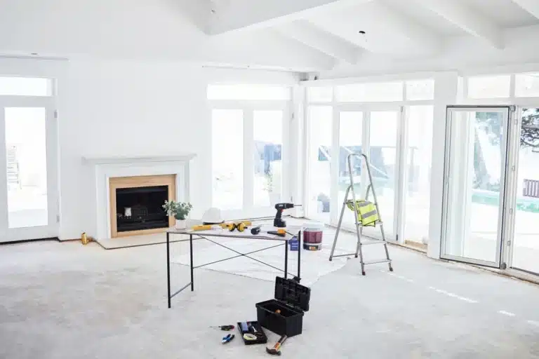 Une pièce lumineuse et aérée en cours de rénovation, dotée de murs blancs et de grandes fenêtres. Une table supporte des outils, à côté d'une échelle et d'une boîte à outils, évoquant un chantier en cours.