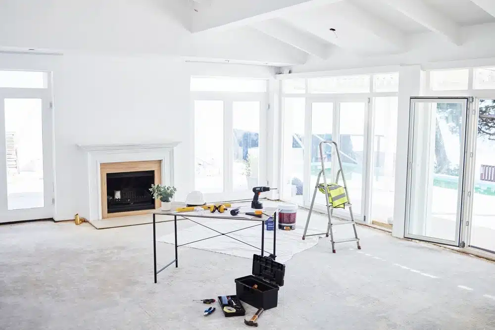 Une pièce lumineuse et aérée en cours de rénovation, dotée de murs blancs et de grandes fenêtres. Une table supporte des outils, à côté d'une échelle et d'une boîte à outils, évoquant un chantier en cours.