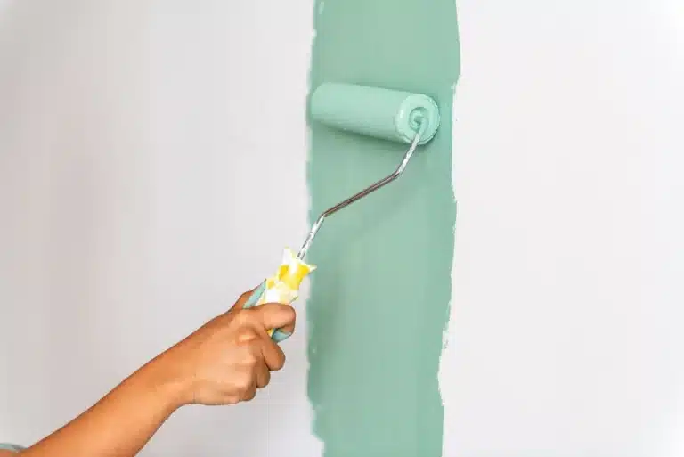 Une main utilise un rouleau pour appliquer de la peinture vert menthe sur un mur blanc, créant une atmosphère fraîche et éclatante. Le rouleau est partiellement recouvert de peinture.