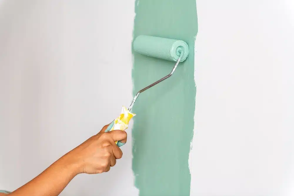 Une main utilise un rouleau pour appliquer de la peinture vert menthe sur un mur blanc, créant une atmosphère fraîche et éclatante. Le rouleau est partiellement recouvert de peinture.