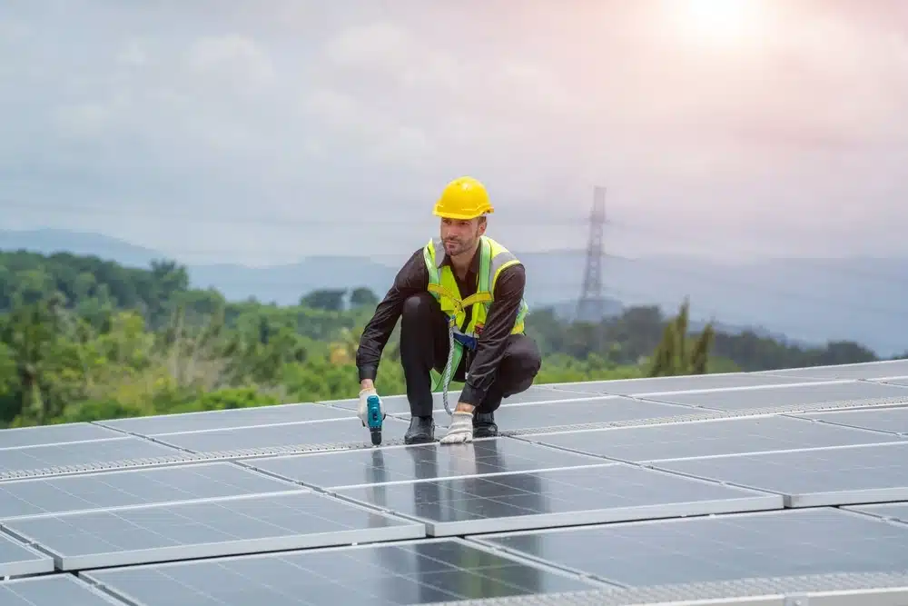 Un ouvrier portant un casque de chantier jaune et un gilet de sécurité installe des panneaux solaires sur un toit, au milieu d'un paysage pittoresque, sous un ciel nuageux traversé par la lumière du soleil.