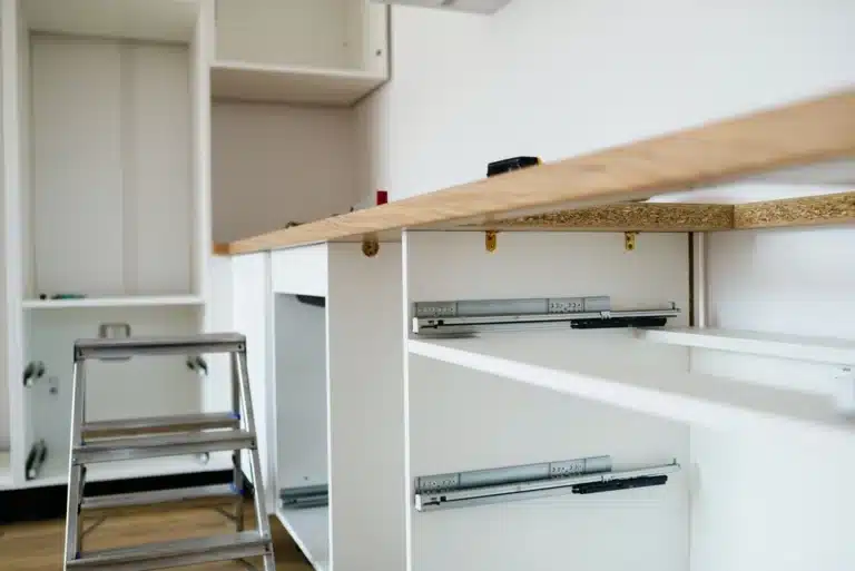Une cuisine en cours de montage présente des placards blancs aux coulisses de tiroir métalliques apparentes ainsi qu'un plan de travail en bois. Un petit escabeau est posé à proximité sur le sol en bois, témoignant de travaux d'installation en cours.