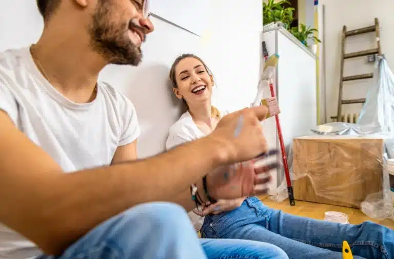Un couple souriant, en tenue décontractée, est assis par terre en train de peindre une pièce. La présence d'une échelle, de meubles recouverts et de matériel de peinture évoque un projet de bricolage joyeux.