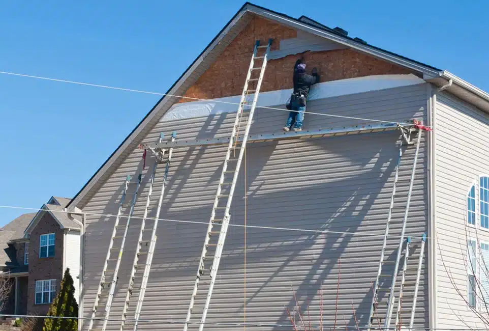 Un ouvrier du bâtiment pose un revêtement extérieur sur la partie supérieure du mur d'une maison, à l'aide d'échelles et d'un échafaudage. Le ciel bleu limpide laisse présager une journée ensoleillée.