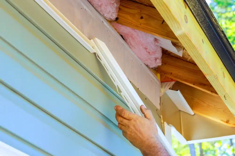 Gros plan sur les mains d'une personne installant un bardage en vinyle blanc sur une maison, avec de l'isolant rose visible. La scène témoigne d'un travail de construction minutieux.