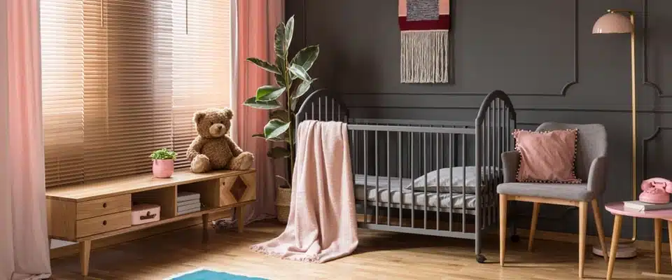 Une chambre de bébé chaleureuse avec un berceau gris, une couverture rose et un ours en peluche posé sur une console en bois. Des rideaux rose tendre et un fauteuil apportent chaleur et charme.