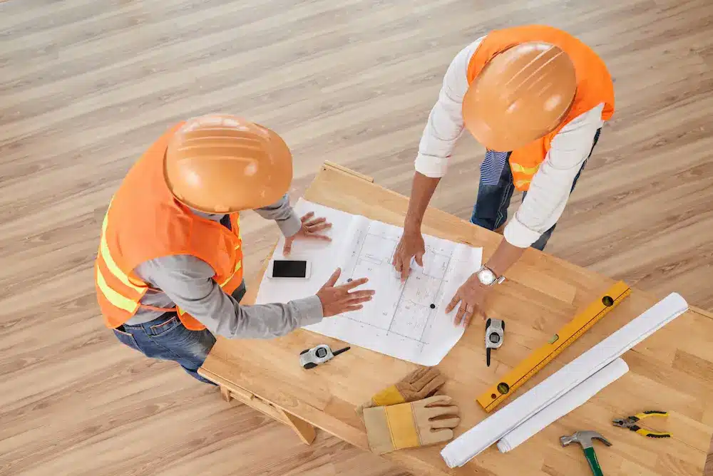 Deux ouvriers du bâtiment, vêtus de gilets et de casques de sécurité orange, examinent des plans sur une table en bois, entourés d'outils comme un niveau et un mètre ruban.