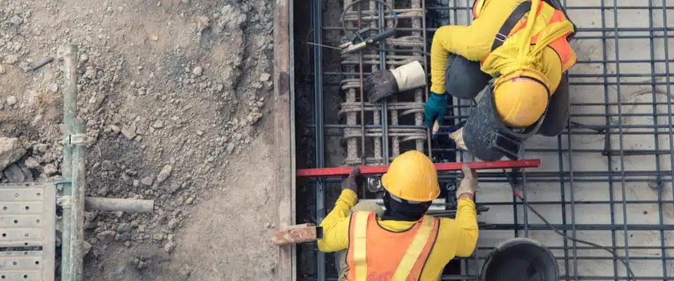 Deux ouvriers du bâtiment, vêtus d'équipements de sécurité jaunes et portant des casques, travaillent sur un treillis de barres d'armature en acier sur un chantier, très concentrés sur leur tâche.