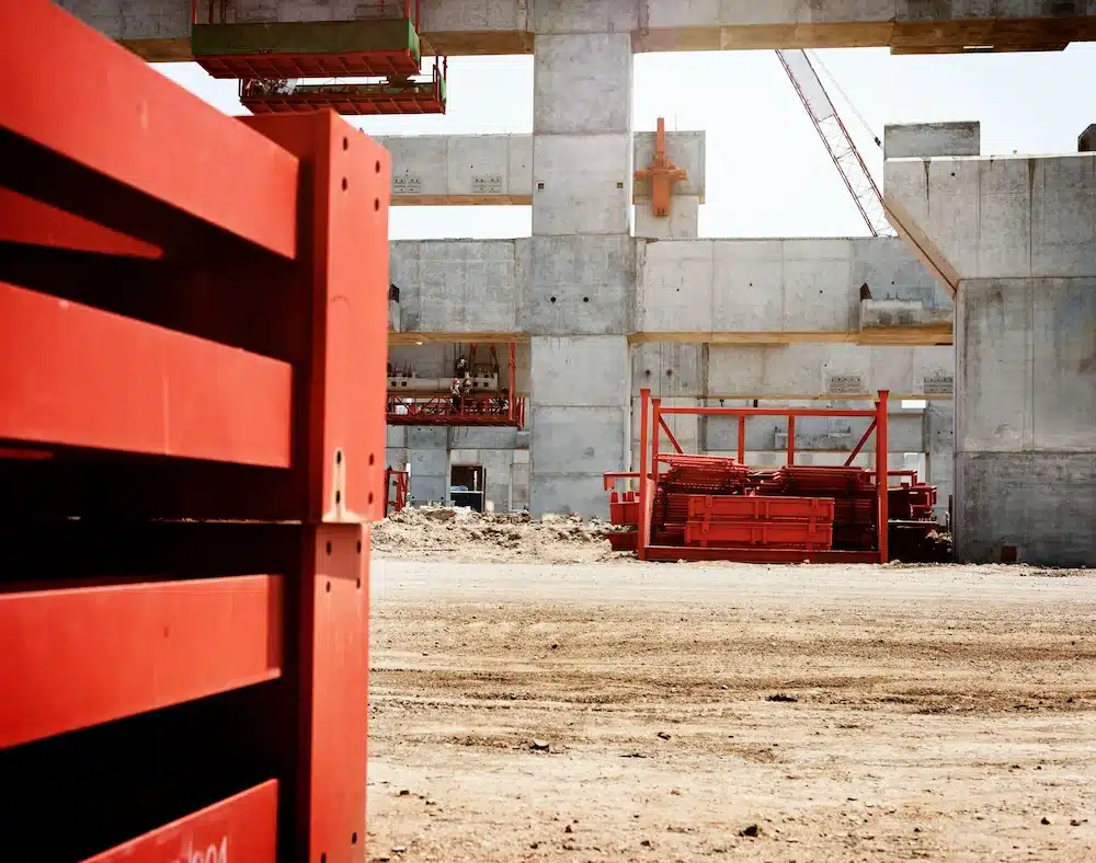 Chantier de construction présentant des structures en béton et des équipements en métal rouge. La scène évoque l'activité laborieuse et le progrès sous une lumière vive.