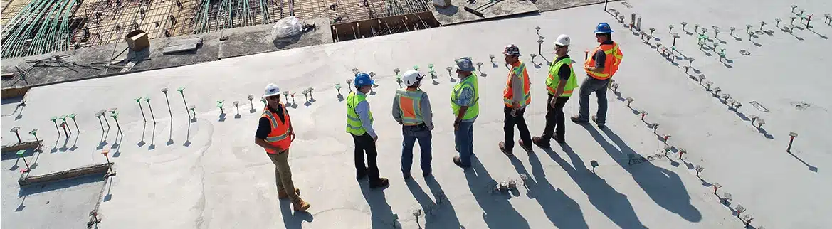 Des ouvriers du bâtiment, portant gilets et casques de sécurité, sont alignés sur un chantier en béton et discutent. Des barres d'armature et du matériel sont visibles en arrière-plan.