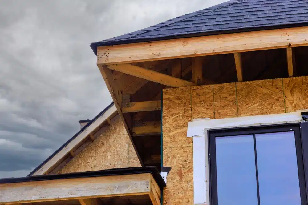 Extérieur d'une maison en cours de construction, avec un bardage en bois et une fenêtre à cadre noir sous un ciel nuageux. La scène présente un aspect brut et inachevé.
