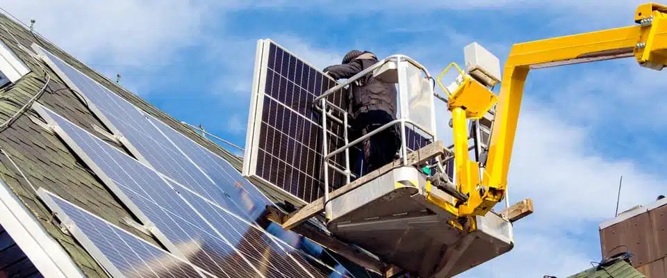 Un ouvrier, installé dans une nacelle élévatrice, installe des panneaux solaires sur un toit en pente sous un ciel bleu limpide. La scène évoque le progrès et la durabilité.