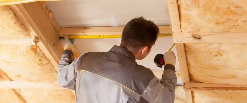 Un homme vêtu d'une veste de travail grise mesure des poutres en bois dans un grenier à l'aide d'un mètre ruban. L'espace est entouré d'un matériau isolant jaune.