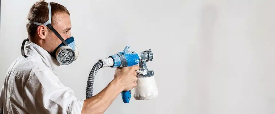 Un homme portant un masque de protection utilise un pistolet pulvérisateur de peinture bleue sur un mur blanc, témoignant de sa concentration et de son professionnalisme dans ce travail de peinture.