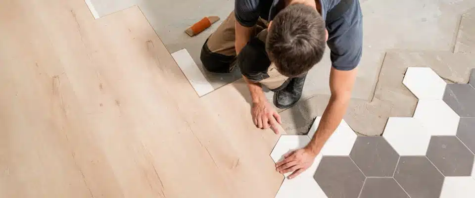 Une personne procède à la pose d'un revêtement de sol, assurant la transition entre des carreaux hexagonaux et des lames de bois. La scène se déroule dans un espace de travail calme, mettant en valeur savoir-faire et précision.