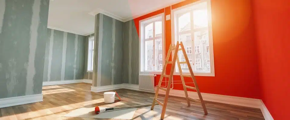 Pièce baignée de lumière avec un escabeau, un seau de peinture et un rouleau posés au sol. Les murs gris et orange vif contrastent, créant une atmosphère dynamique.