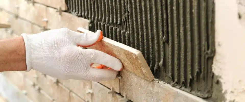 Une main gantée applique un carreau de céramique sur un mur préalablement encollé. L'image traduit la concentration et la précision du processus de pose.