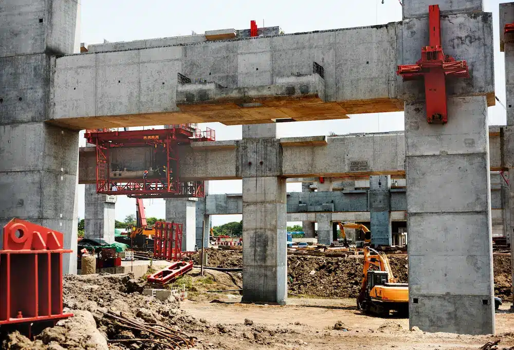 Chantier de construction d'un pont en béton avec grues, échafaudages et engins lourds. De grands piliers et des poutres dominent la scène, témoignant de l'avancement des travaux.